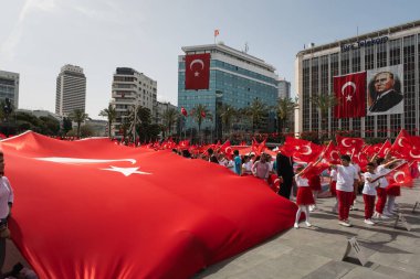 İzmir, Türkiye - 23 Nisan 2024: Ulusal Egemenlik ve Çocuk Günü sırasında, çocuklar İzmir Cumhuriyet Meydanı 'na dev bir Türk bayrağı gererken, daha fazla katılımcı, dev bir bayrak ve arkasında Atatürk portresi görülebiliyor..