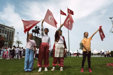 İzmir, Türkiye 23 Nisan 2024 Çocuklar Günü kutlamalarında dört neşeli kız geleneksel gösteri elbiseleriyle Türk bayrağını sallarken, arka planda at sırtında Atatürk heykeli