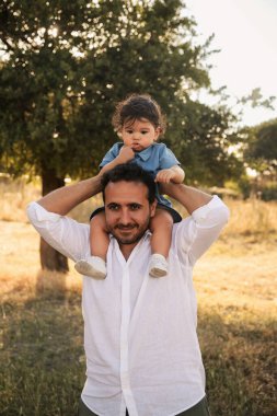 Bir yaşındaki bir çocuk, arka planda ağaçlar ve bokeh ile babasının omuzlarında oturuyor ve Babalar Günü 'nde özel bir anı yakalıyor.