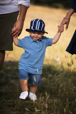 Bir yaşında bir çocuk doğada yürüyor, ailesinin elini tutuyor, mavi bir şapka, mavi gömlek ve mavi kot pantolon giyiyor..
