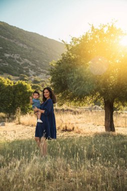 Mavi elbiseli bir Özbek anne, bir yaşındaki oğlunu arka planda bir ağaçla çimenli doğal bir ortamda tutuyor.