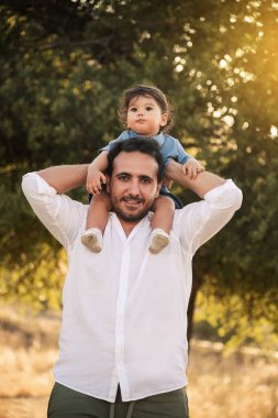 Bir yaşındaki bir çocuk, arka planda ağaçlar ve bokeh ile babasının omuzlarında oturuyor ve Babalar Günü 'nde özel bir anı yakalıyor.