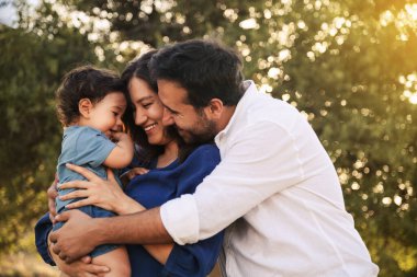 Mavi elbiseli bir baba, mavi elbiseli bir anne ve arka planda bir ağaçla çimenli doğal bir ortamda sarılıp gülümseyen bir oğullarıyla neşeli bir aile anı.
