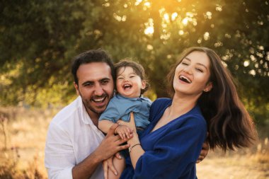 Mavi elbiseli bir baba, mavi elbiseli bir anne ve arka planda bir ağaçla çimenli doğal bir ortamda sarılıp gülümseyen bir oğullarıyla neşeli bir aile anı.