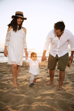 Beyaz gömlekli ve güneş gözlüklü bir çocuk plajda ailesinin elini tutarak yürüyor. Arka planda kum ve gün batımının ılık ışığı var.