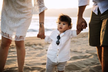 Beyaz gömlekli ve güneş gözlüklü bir çocuk plajda ailesinin elini tutarak yürüyor. Arka planda kum ve gün batımının ılık ışığı var.