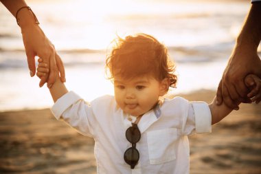 Beyaz gömlekli ve güneş gözlüklü bir çocuk plajda ailesinin elini tutarak yürüyor. Arka planda kum ve gün batımının ılık ışığı var.