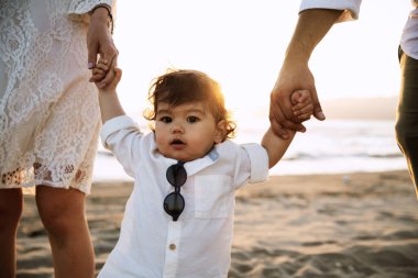 Beyaz gömlekli ve güneş gözlüklü bir çocuk plajda ailesinin elini tutarak yürüyor. Arka planda kum ve gün batımının ılık ışığı var.