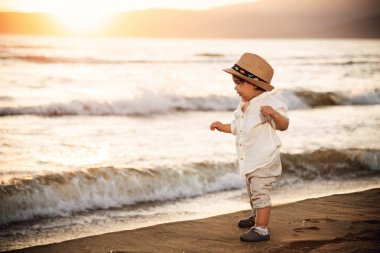 Beyaz gömlekli ve şapkalı bir çocuk gün batımında plajda koşuyor. Arka planda deniz ve güzel bir günbatımı var.