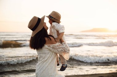 Beyaz elbiseli ve şapkalı neşeli bir anne gün batımında beyaz gömlekli ve şapkalı oğlunu kumsalda tutuyor. Her ikisi de gülümsüyor, arkada deniz ve gün batımı var..