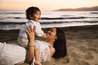 Bir yaşında beyaz gömlekli bir çocuk plajda annesinin üzerine oturuyor. İkisi de gülümsüyor ve arkada deniz ve günbatımından mutlular.
