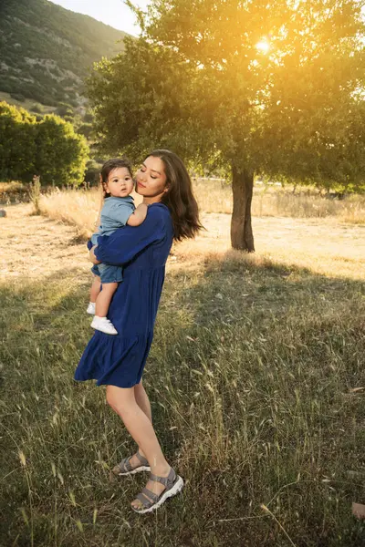 stock image An Uzbek mother in a blue dress holding her one-year-old son in a grassy natural environment with a tree in the background