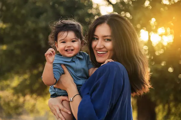 stock image An Uzbek mother in a blue dress holding her one-year-old son in a grassy natural environment with a tree in the background