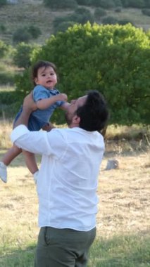 Beyaz gömlekli bir baba, bir yaşındaki oğlunu çimenli bir doğal ortamda neşeyle havaya kaldırıyor. Bir ağaç ve çimenlik bir alan arkaplan oluşturur.