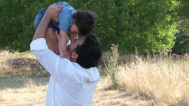 Beyaz gömlekli bir baba, bir yaşındaki oğlunu çimenli bir doğal ortamda neşeyle havaya kaldırıyor. Bir ağaç ve çimenlik bir alan arkaplan oluşturur.