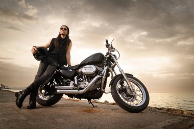 30 'lu yaşlarında metal elbiseli bir adam gün batımında arkasında manzaralı bir okyanus manzarası olan motosikletine yaslanıyor..