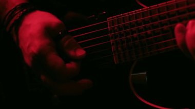 Kırmızı ışık altında ustalıkla gitar çalan bir elin yakın plan videosu ayrıntılı parmak hareketlerini ve dramatik atmosferi vurguluyor