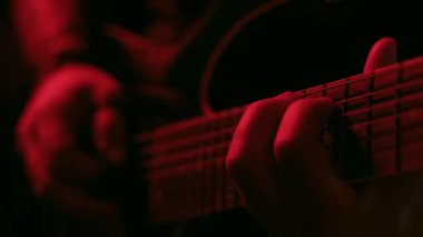 Kırmızı ışık altında ustalıkla gitar çalan bir elin yakın plan videosu ayrıntılı parmak hareketlerini ve dramatik atmosferi vurguluyor