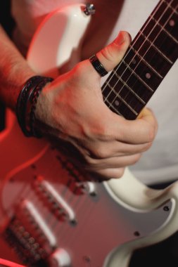 Beyaz gitarı kavrayan bir elektro gitaristin, hem normal hem de kırmızı ışıkla aydınlatılan, enstrümanı ve müzisyenin tarzını gösteren detaylı bir yakın çekimi.