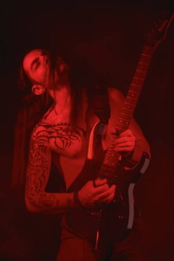 Ayakta dururken elektro gitar çalan uzun saçlı bir metal gitaristin portresi dramatik bir etki yaratmak için kırmızı ışıkla aydınlatılmış.