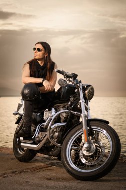 30 'lu yaşlarda metal elbiseli bir adam gün batımında arkasında manzaralı bir deniz manzarası olan motosikletinin üzerinde oturuyor..