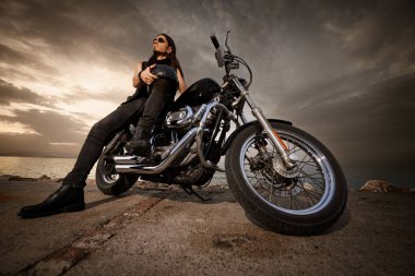 30 'lu yaşlarda metal elbiseli bir adam gün batımında arkasında manzaralı bir deniz manzarası olan motosikletinin üzerinde oturuyor..