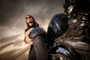 Rock ya da metal kıyafetli bir adam motosikletinin üzerinde oturur ve arkasında dramatik bir gökyüzü ile poz verir. Miğferini dizine takmış ve soğukkanlı ve kendinden emin bir tavır sergiliyor..