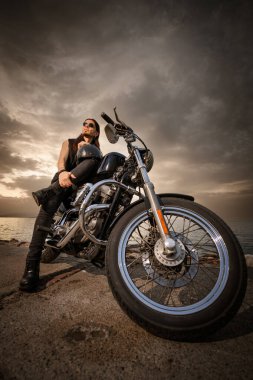 30 'lu yaşlarda metal elbiseli bir adam gün batımında arkasında manzaralı bir deniz manzarası olan motosikletinin üzerinde oturuyor..