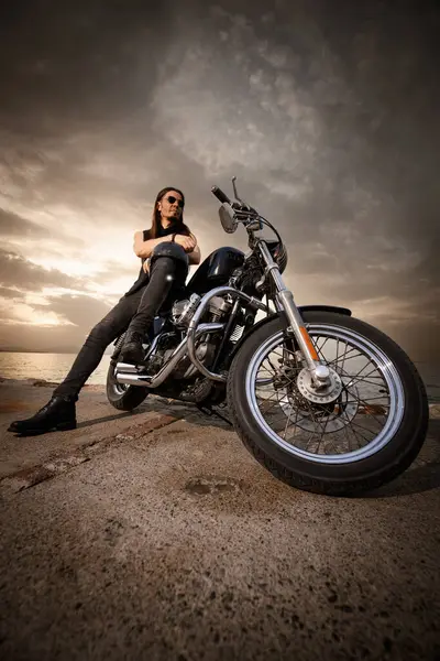 30 'lu yaşlarda metal elbiseli bir adam gün batımında arkasında manzaralı bir deniz manzarası olan motosikletinin üzerinde oturuyor..