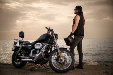 Kaya gibi giyinmiş bir motorcu, sahil kenarında park etmiş motosikletinin yanında kaskını tutuyor. Sahne, sakin okyanus manzarasını, özgürlük ve macera hissini yakalar.