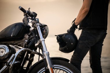 Arka planda deniz ve gökyüzü olan helikopter stili motosikletinin yanında kaskını tutan motorcunun yakın plan fotoğrafı. Görüntü, özgürlük ve macera hissini yakalar.