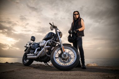 Arka planda deniz ve gökyüzü olan helikopter stili motosikletinin yanında kaskını tutan motorcunun yakın plan fotoğrafı. Görüntü, özgürlük ve macera hissini yakalar.