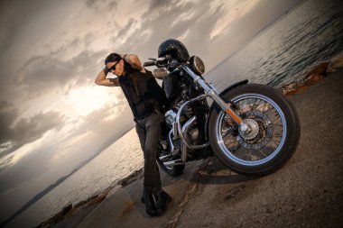 Helikopter stili motosikletine yaslanmış uzun saçlı bir motorcu deniz kenarında saçını topluyor gibi görünüyor. Deri bir kask motosiklet koltuğunda duruyor, engebeli sahneyi büyütüyor.