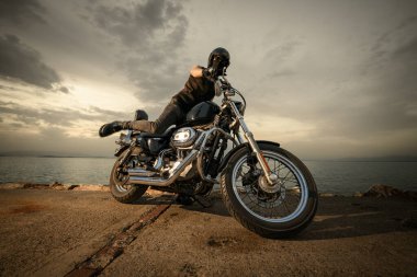 Kaskı, gözlükleri ve yüz örtüsüyle donatılmış bir motorcu bulutlu bir günbatımında deniz kenarında bir motosikletin üzerinde oturuyor. Görüntü bir macera ve özgürlük hissi yakalıyor.