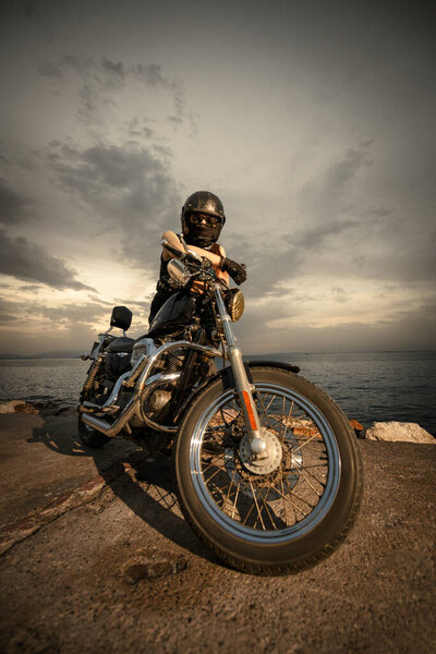 A biker fully geared with a helmet, goggles, and face covering, sitting on a chopper motorcycle by the sea during a cloudy sunset. The image captures a sense of adventure and freedom