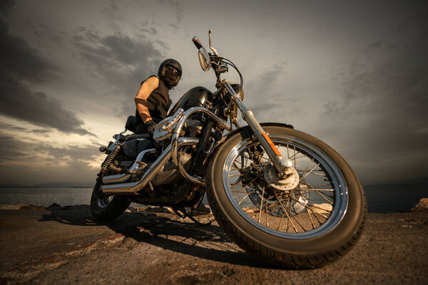 A biker fully geared with a helmet, goggles, and face covering, sitting on a chopper motorcycle by the sea during a cloudy sunset. The image captures a sense of adventure and freedom