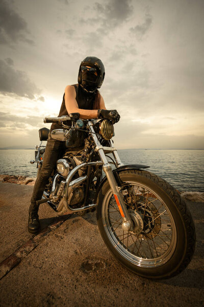 A biker fully geared with a helmet, goggles, and face covering, sitting on a chopper motorcycle by the sea during a cloudy sunset. The image captures a sense of adventure and freedom