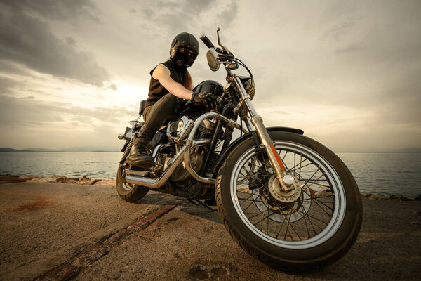 A biker fully geared with a helmet, goggles, and face covering, sitting on a chopper motorcycle by the sea during a cloudy sunset. The image captures a sense of adventure and freedom