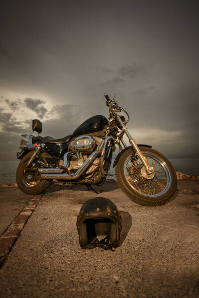 A chopper-style motorcycle stands by the seaside with a leather helmet placed on the ground. The cloudy sky and calm sea set a peaceful scene as the sun has set
