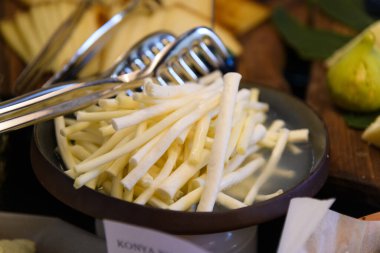 Otelin açık büfesinde sergilenen çeşitli peynirli çubukların yakın plan çekimi. Yanında da servis için metal maşalar var.