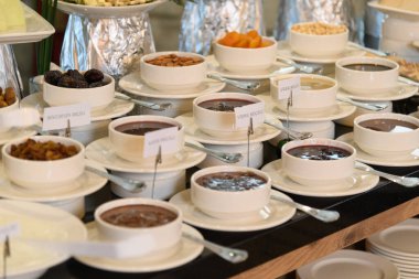 Çilek, kiraz, böğürtlen gibi çeşitli reçel çeşitlerinin yakın çekimi bir otel kahvaltı büfesinde, etiketli beyaz kaselerde servis edilir.