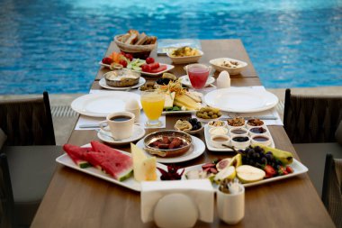 Havuzun kenarına yayılmış müsrif bir kahvaltı, çeşitli meyveler, zeytinler, reçeller ve ekmekler, mükemmel bir sabah ortamı yaratıyor.