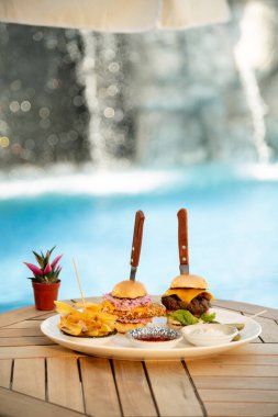 Havuz kenarındaki tabakta iki hamburger, patates kızartması ve sosla yakın plan. Rahatlatıcı bir arkaplanı olan rahatlatıcı bir öğle yemeği için ideal.