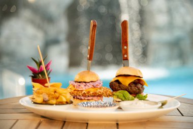 Havuz kenarındaki tabakta iki hamburger, patates kızartması ve sosla yakın plan. Rahatlatıcı bir arkaplanı olan rahatlatıcı bir öğle yemeği için ideal.