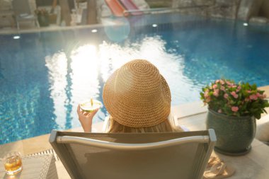 Şapkalı ve mayo giyen bir kadın havuz kenarında rahatlar, derginin tadını çıkarır ve suna balığında ferahlatıcı bir kokteyl içer.