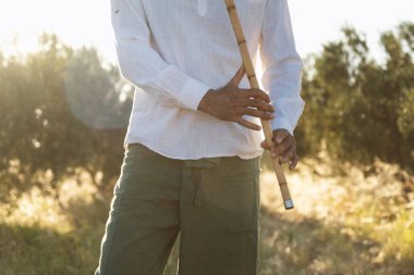 Zeytin tarlasının huzurlu atmosferiyle çevrili, arka planda parlayan güneş ışığıyla çevrili bir adamın el ele tutuşup bir ney flütü çalması.