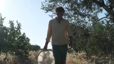 Bir Orta Doğulu adam zeytin koruluğunda yürüyor, elinde bir çerçeve varili tutuyor güneş ışığı ağaçların arasından süzülüyor, huzurlu ve huzurlu bir atmosfer yaratıyor.