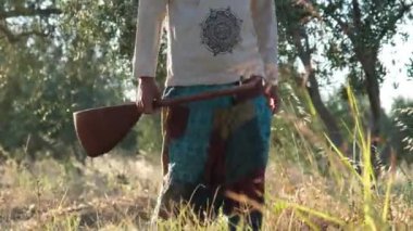 40 'lı yaşlarda bir Orta Doğulu elinde kopuz olan bir zeytin korusundan geçiyor. Güneş ışığı ağaçların arasından süzülürken doğayla olan dinginliği ve bağlantıyı yakalıyor.