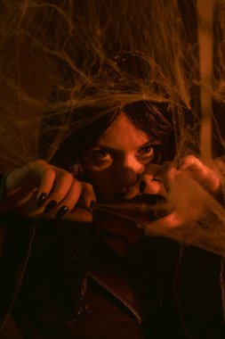 Koyu makyajlı bir cadının yakından çekilmiş portresi. Örümcek ağlarının arasından yoğun bir şekilde bakıyor. Sıcak ışıklandırma ürkütücü, mistik havayı güçlendiriyor. Cadılar Bayramı temalı bir görüntü için mükemmel.