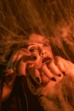 Koyu makyajlı bir cadının yakından çekilmiş portresi. Örümcek ağlarının arasından yoğun bir şekilde bakıyor. Sıcak ışıklandırma ürkütücü, mistik havayı güçlendiriyor. Cadılar Bayramı temalı bir görüntü için mükemmel.